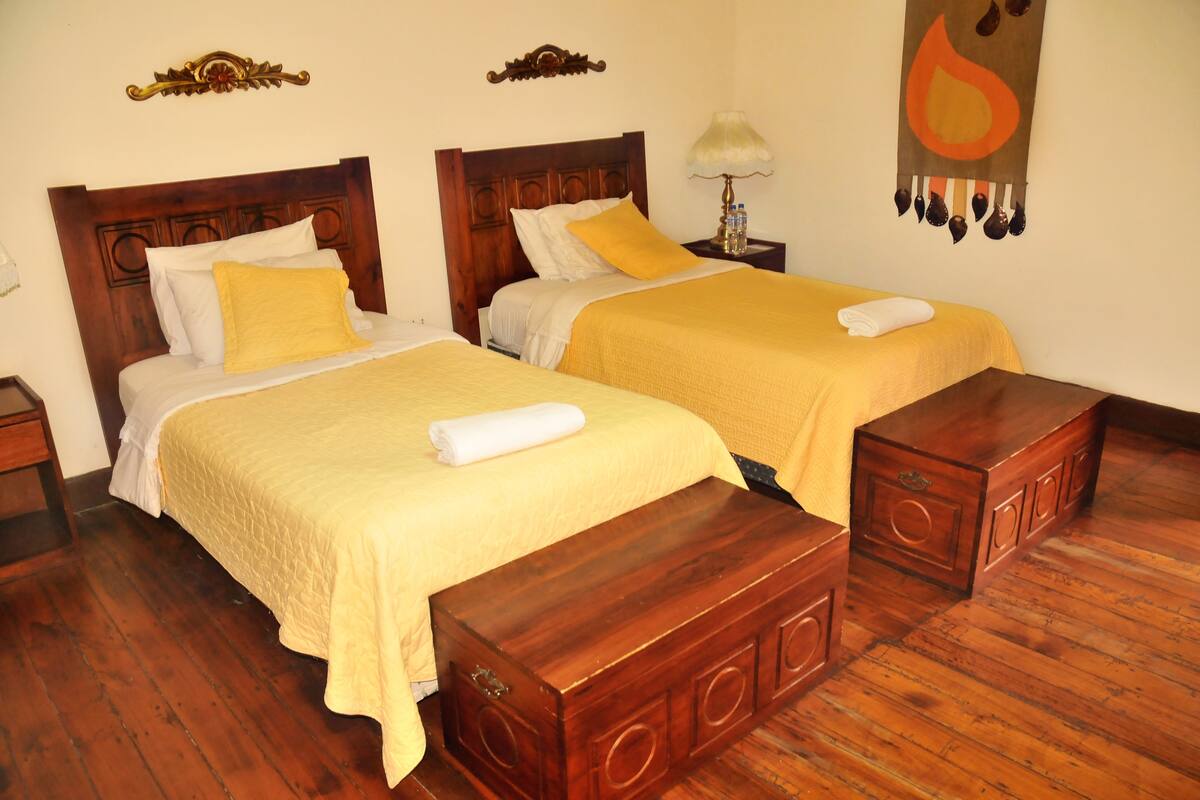 Une chambre d’hôtel avec deux lits en bois, chacun recouvert d’une couvre-lit jaune et agrémenté d’oreillers blancs. On y trouve une table de chevet en bois avec une lampe, ainsi qu’une tapisserie murale.