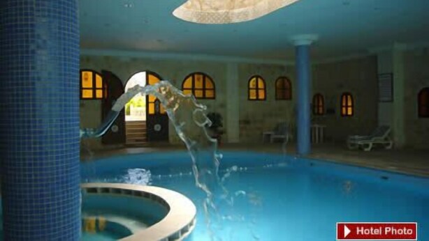 En indendørs swimmingpool med krystalklart vand, en central springvand og buede vinduer i baggrunden.