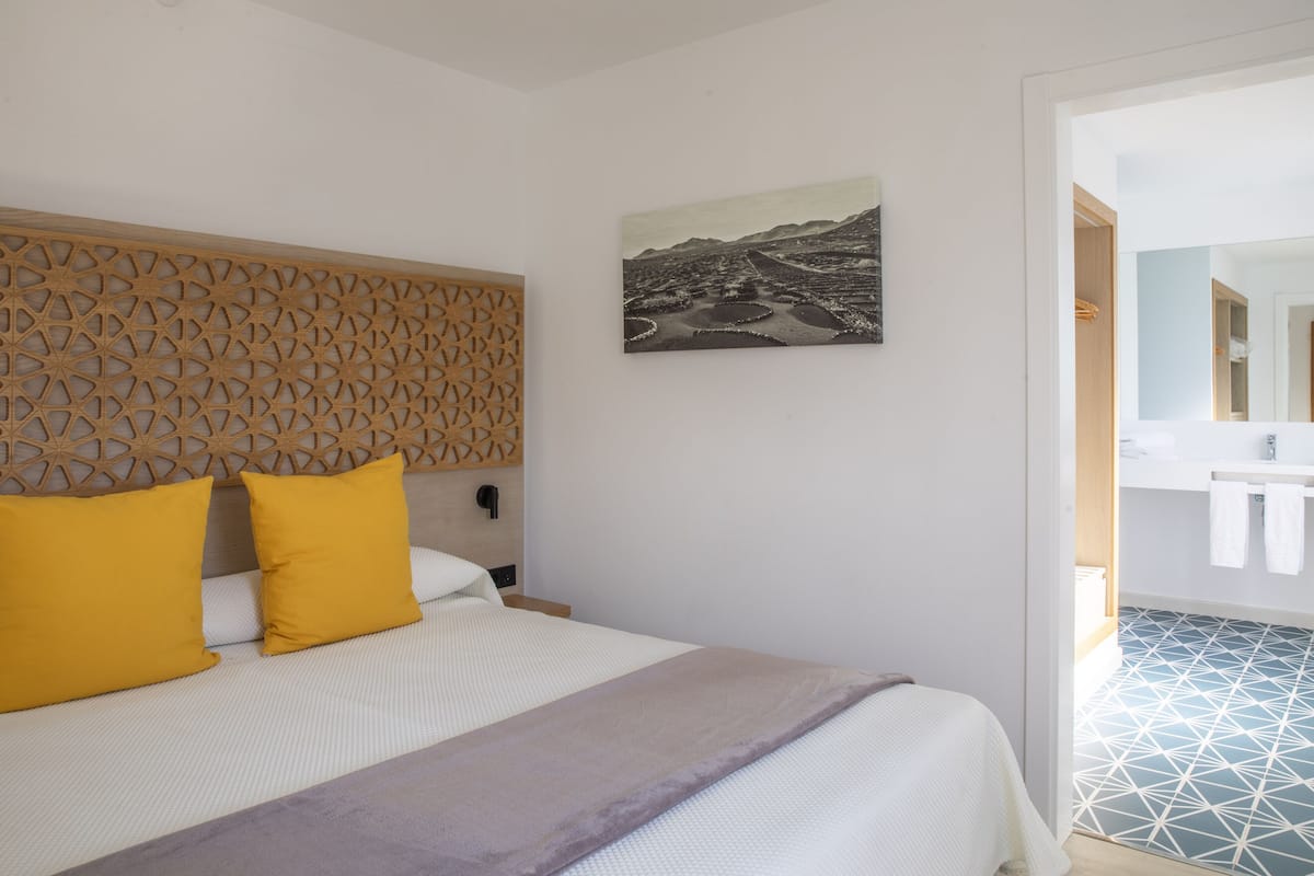 A hotel room with a bed, a wooden headboard, a painting, and a bathroom visible through an open door.