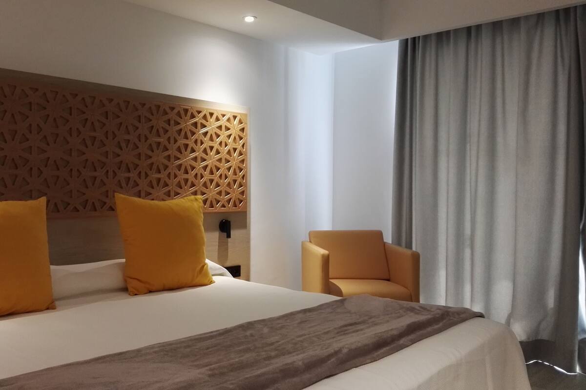A modern hotel room with a large bed, two yellow pillows, a brown throw blanket, a wooden headboard, a side table, and a chair.