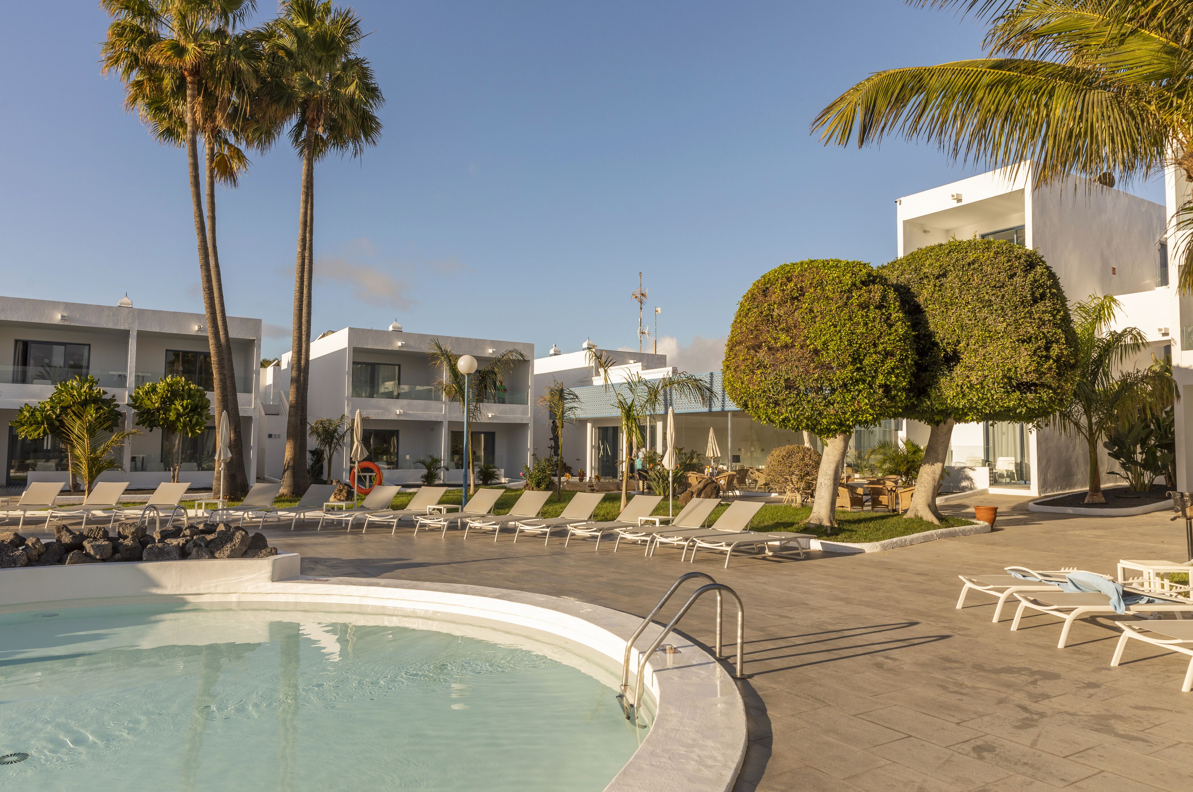 Zona de piscina con sillones de descanso, palmeras y edificios modernos.