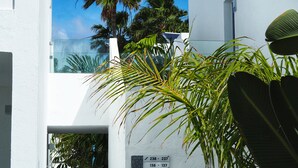 A modern building with a white exterior, a glass balcony, and a palm tree in the foreground. - Oasis Lanz Beach Mate (Teguise)