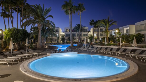 A swimming pool with lounge chairs and palm trees, surrounded by modern buildings. - Oasis Lanz Beach Mate (Teguise)