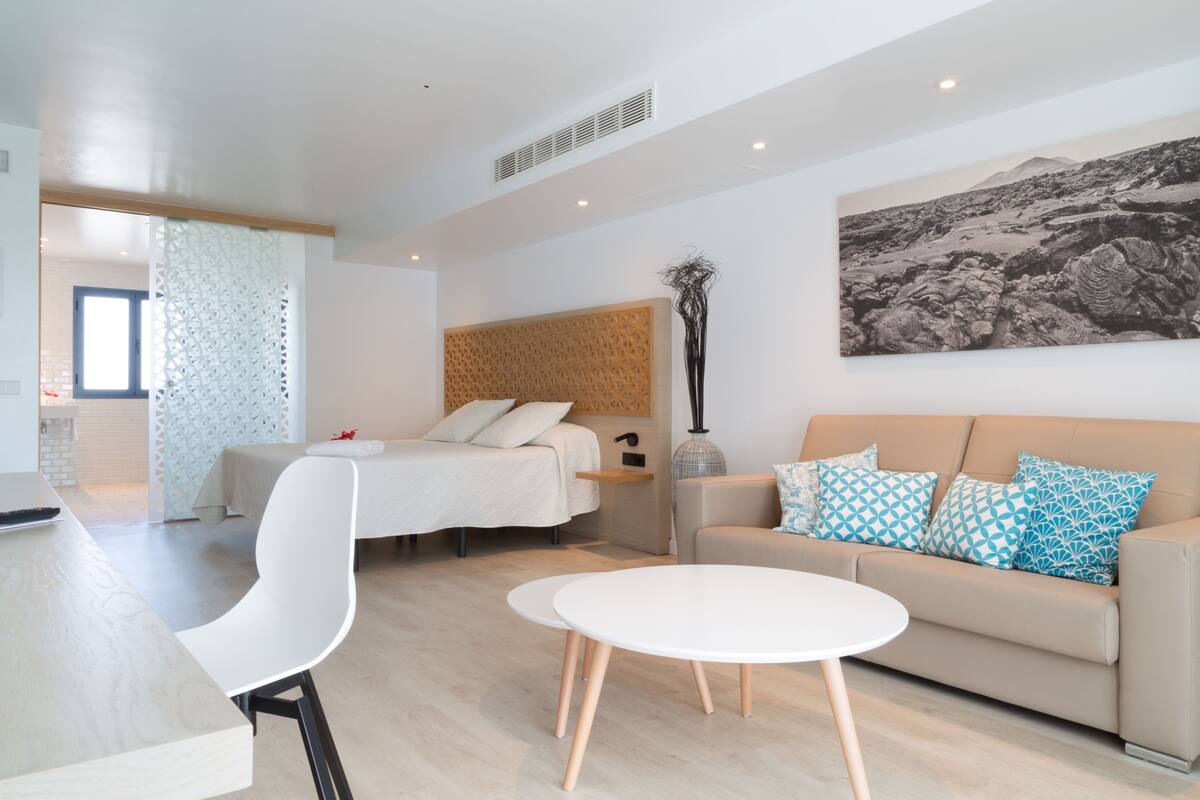 A modern hotel room with a bed, a sofa, a dining table, and a large wall art piece.