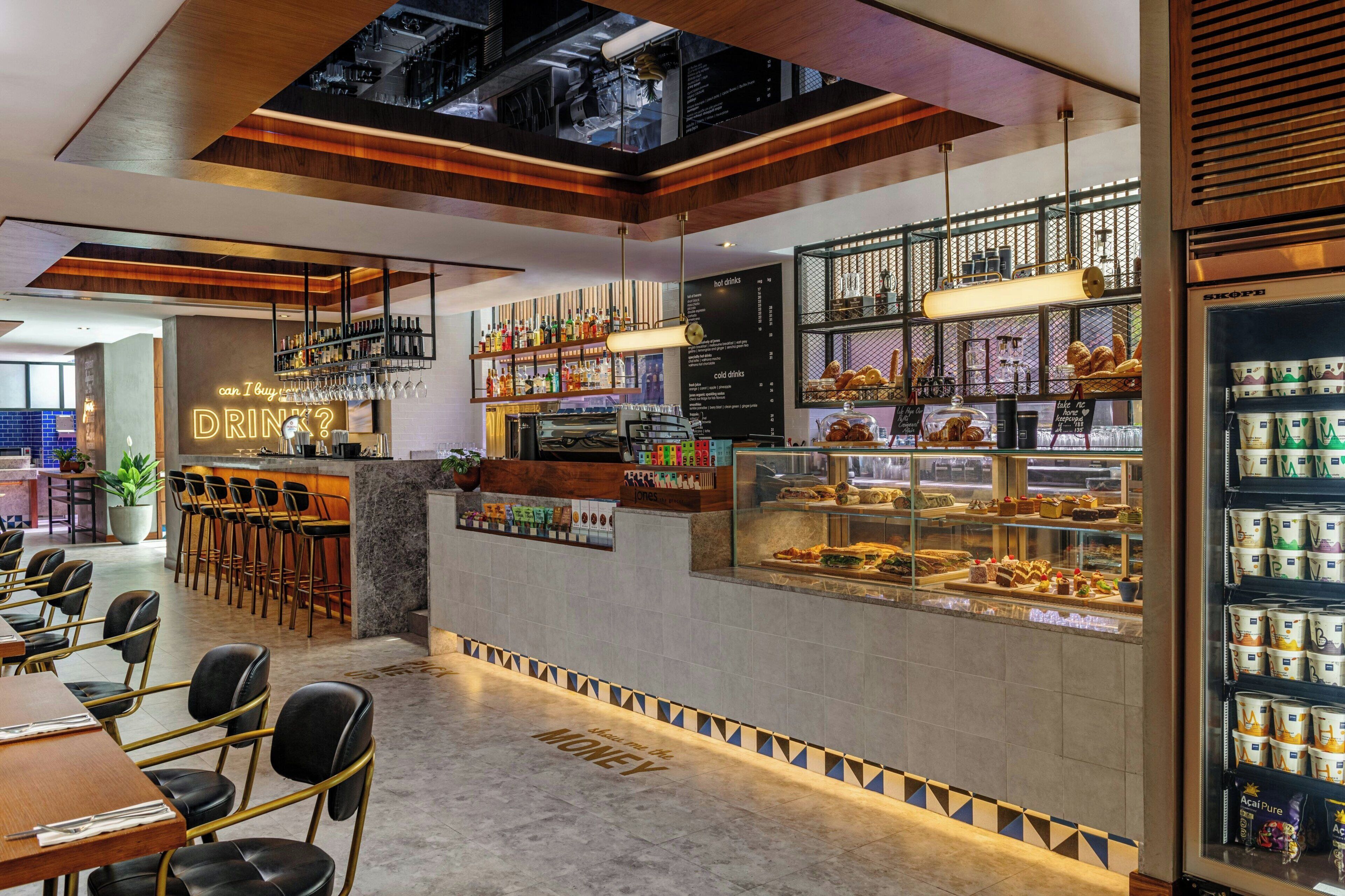 Un espace bar moderne proposant une variété de boissons et de pâtisseries, des tabourets de bar et un coin salon avec des tables et des chaises.