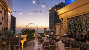 Un bar en la azotea con sillas de mimbre y una barra, con vistas a un paisaje urbano y una noria al atardecer.