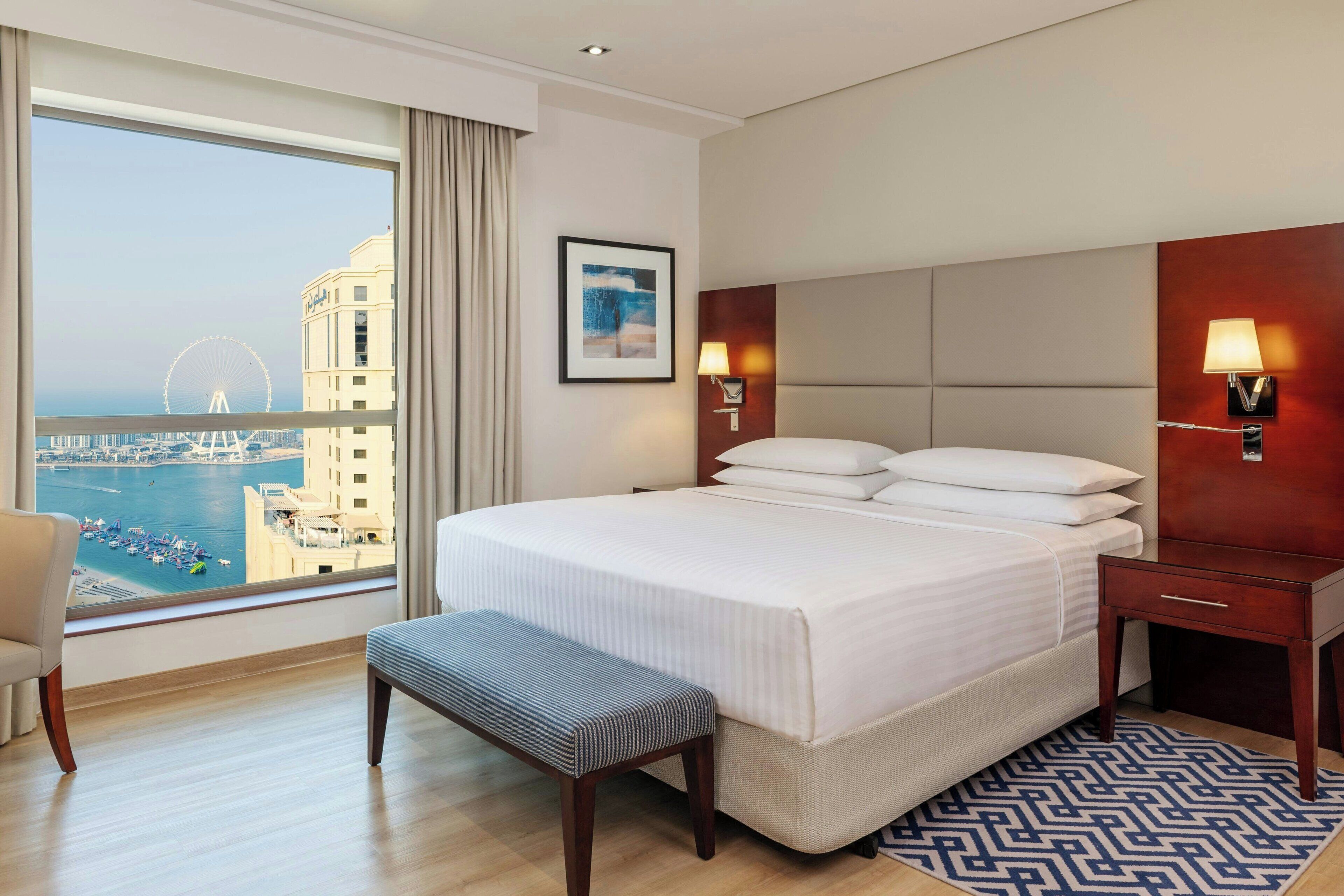 A hotel room with a large bed, a bench, a nightstand, a lamp, and a view of a cityscape and water.