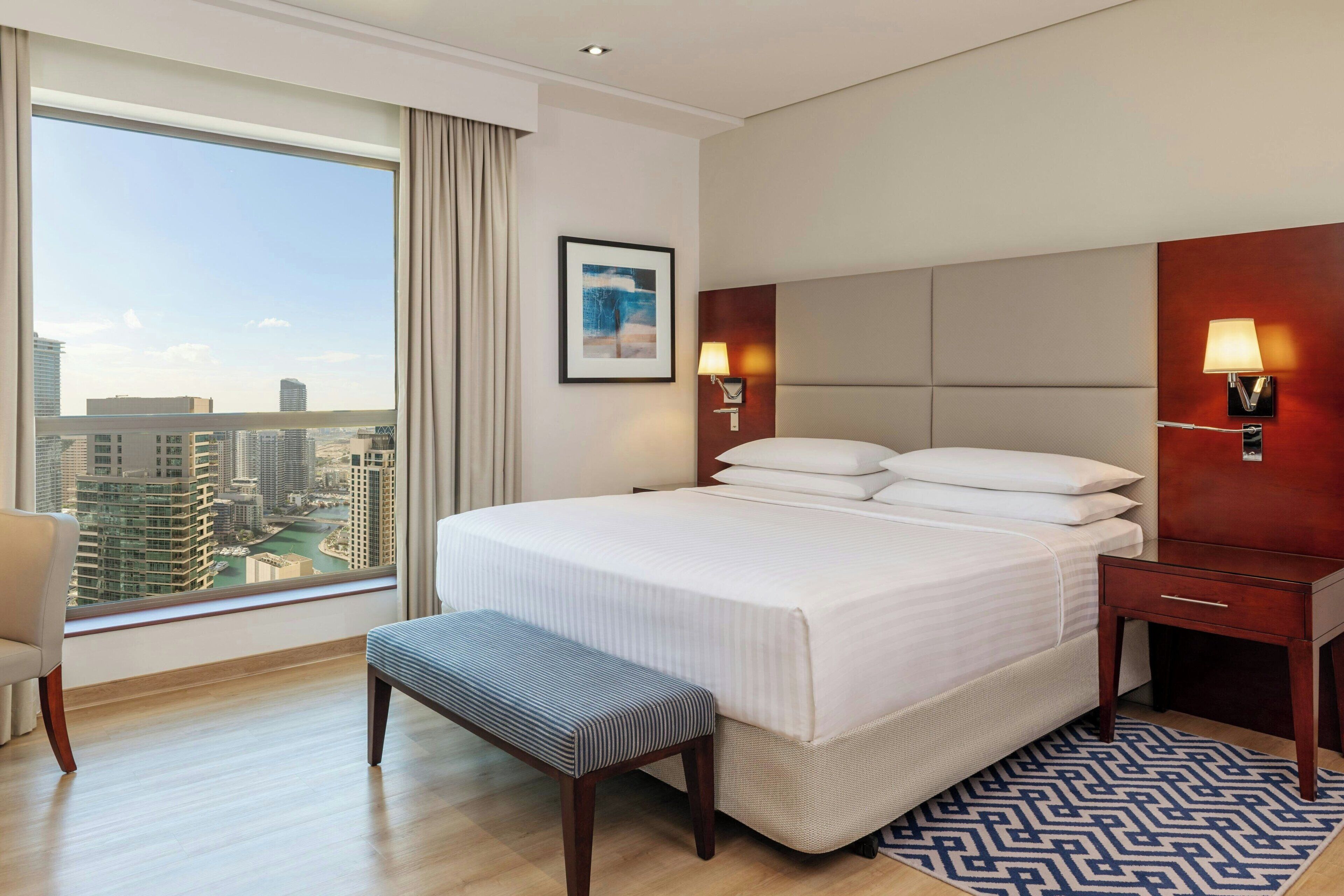 A hotel room with a large bed, a bench, a nightstand, a lamp, and a view of the cityscape.