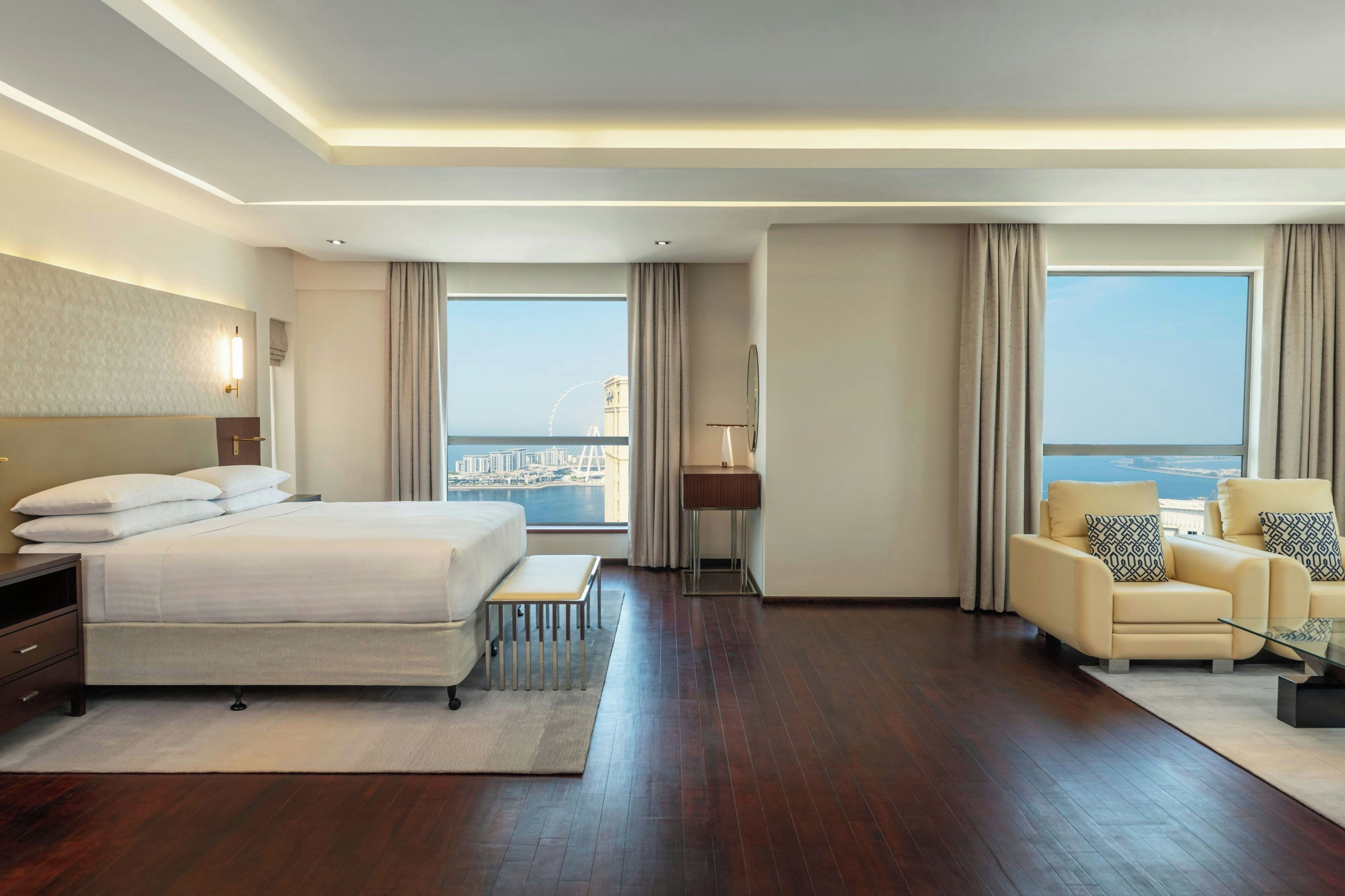 Une chambre d’hôtel comprenant un lit, un coin salon avec un fauteuil, une petite table et une vue sur la ville et l’eau.
