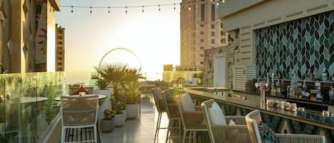 Un bar en la azotea con mesas al aire libre, una noria al fondo y vistas del horizonte de la ciudad.