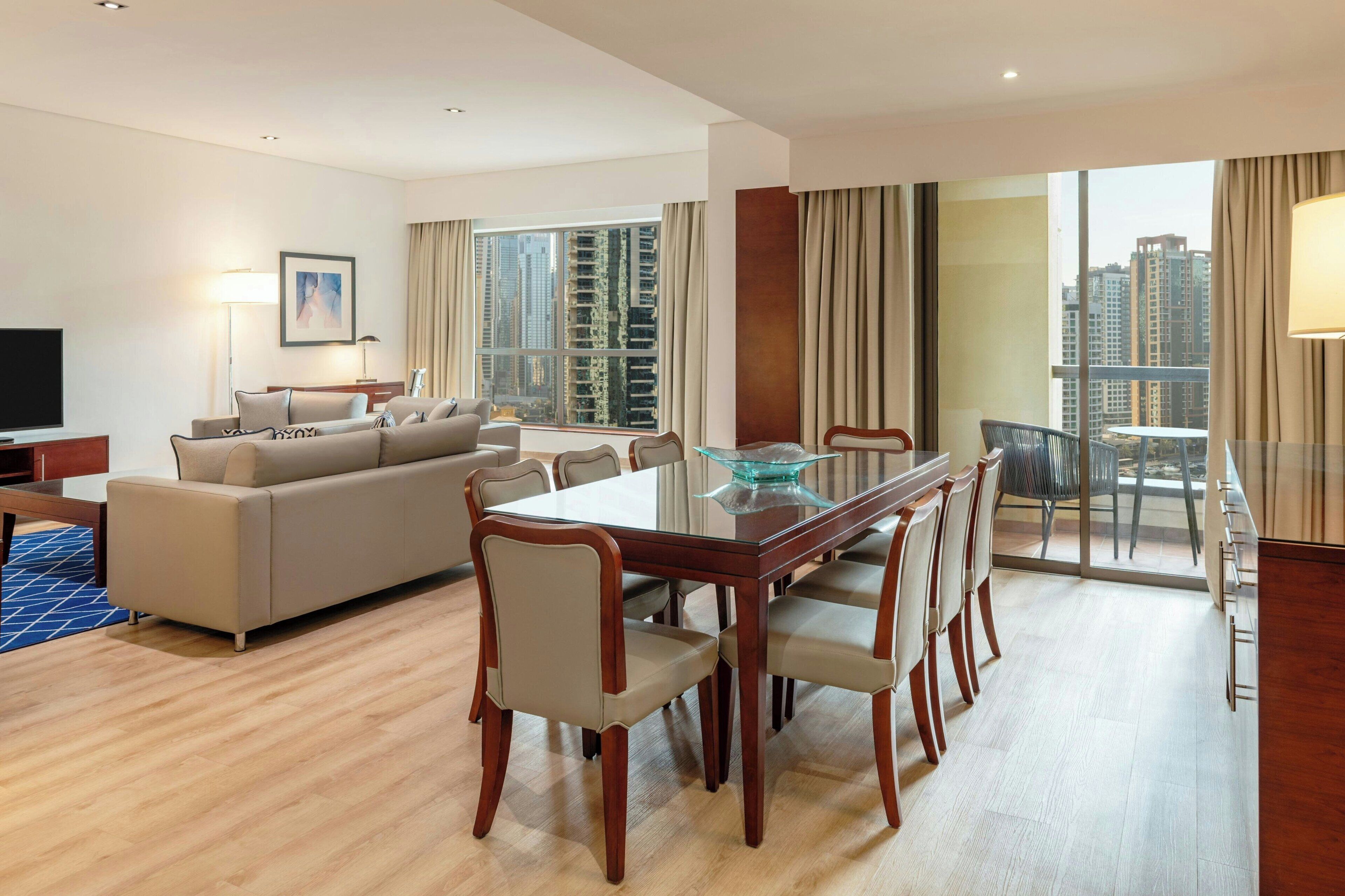 Una habitación de hotel moderna con zona de estar, mesa de comedor y amplios ventanales con vistas a la ciudad.