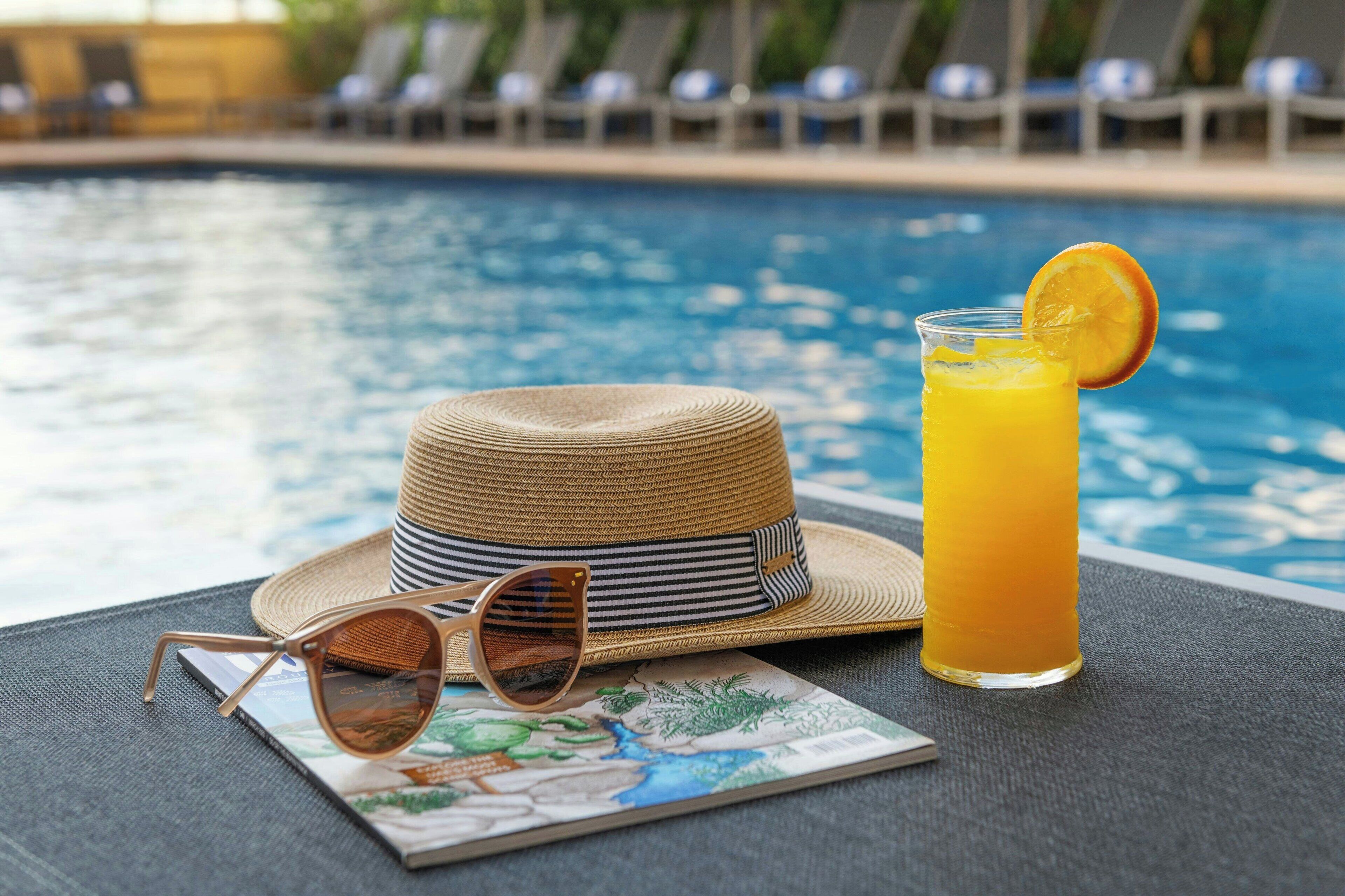 Escena junto a la piscina con un sombrero de paja, gafas de sol y un vaso de zumo de naranja.