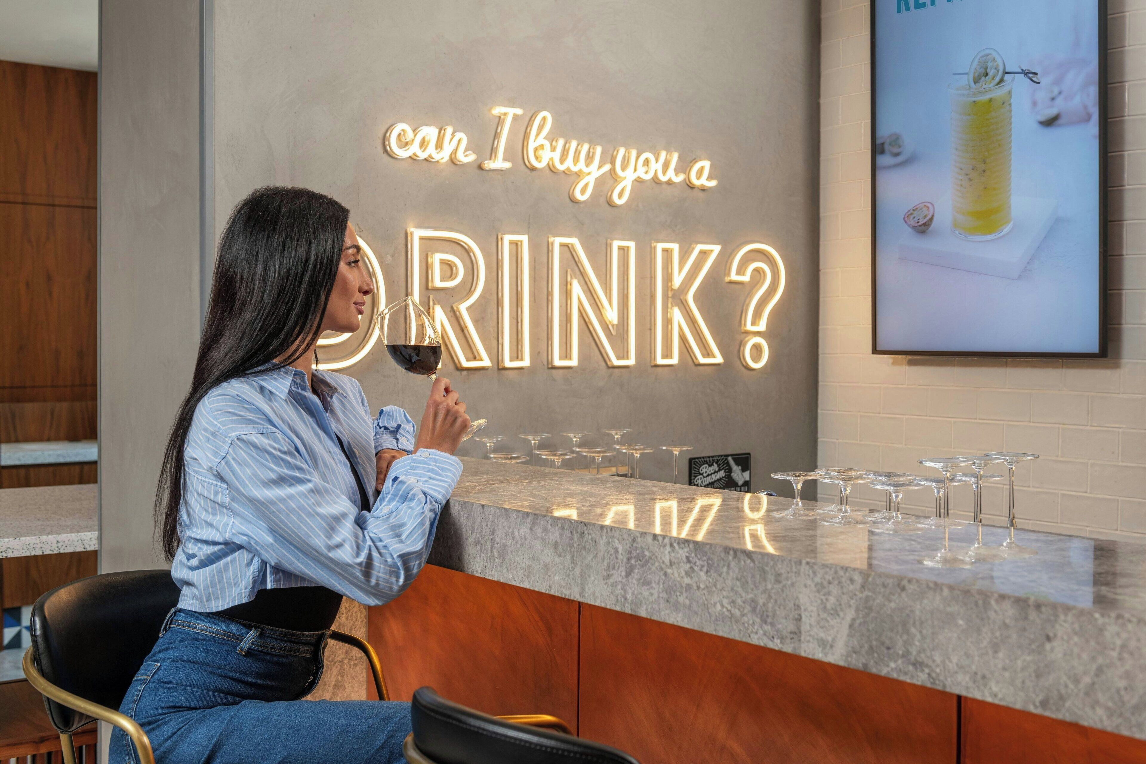 Eine Frau sitzt an einer Bar, im Hintergrund leuchtet ein Neonlicht mit der Aufschrift 'can I buy you a DRINK?', auf der Theke stehen Gläser.