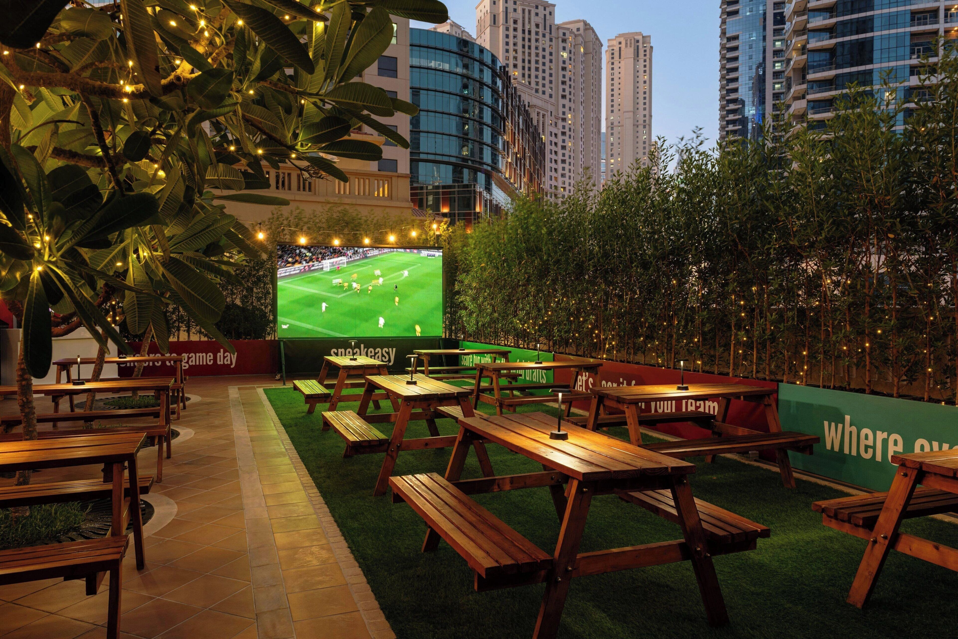 Un espace extérieur aménagé avec des tables et des bancs en bois, un grand écran diffusant un événement sportif, et un décor composé d’immeubles imposants.