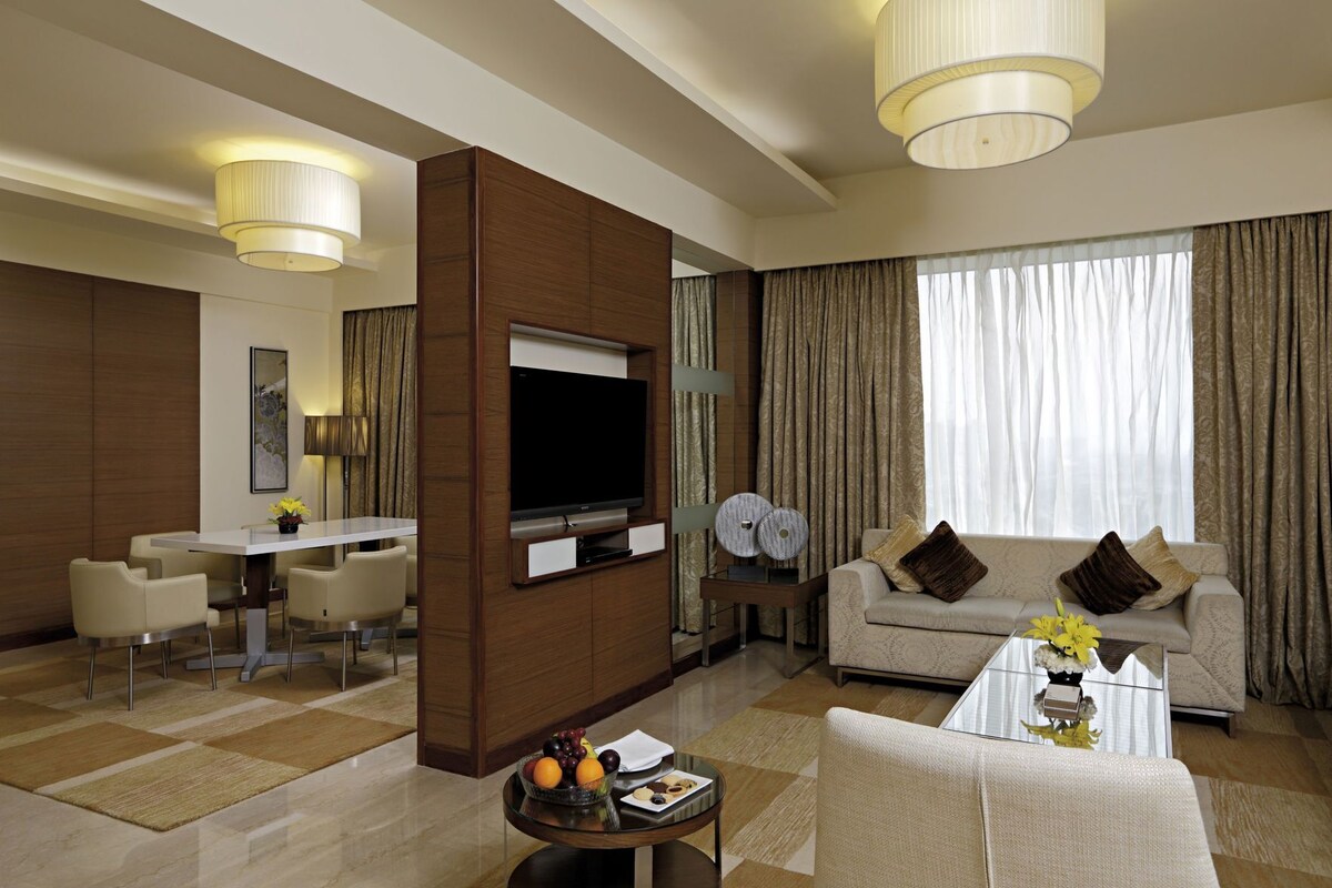 A modern hotel room with a flat-screen TV, a dining area, a sofa, and a coffee table with fruit.