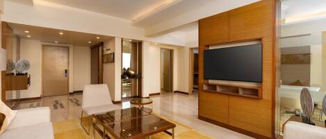 A modern living room with a glass coffee table, white sofas, a flat-screen TV mounted on a wooden wall, and a kitchen area in the background.