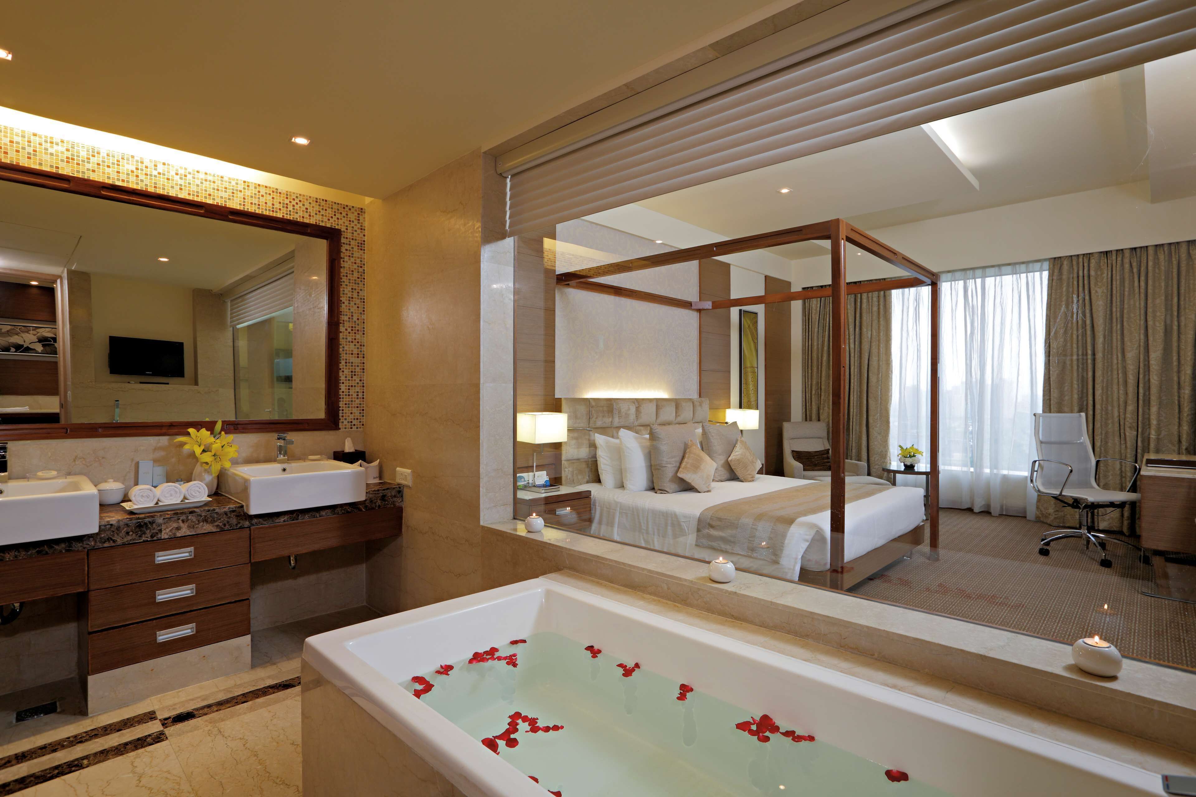 A hotel room with a large mirror, a bathtub with rose petals, a bed with a canopy, and a desk with a chair.
