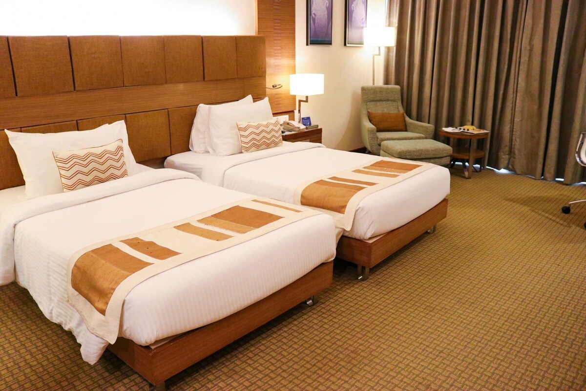 Habitación de hotel con dos camas, cabecera de madera, una silla, una mesita y una lámpara.