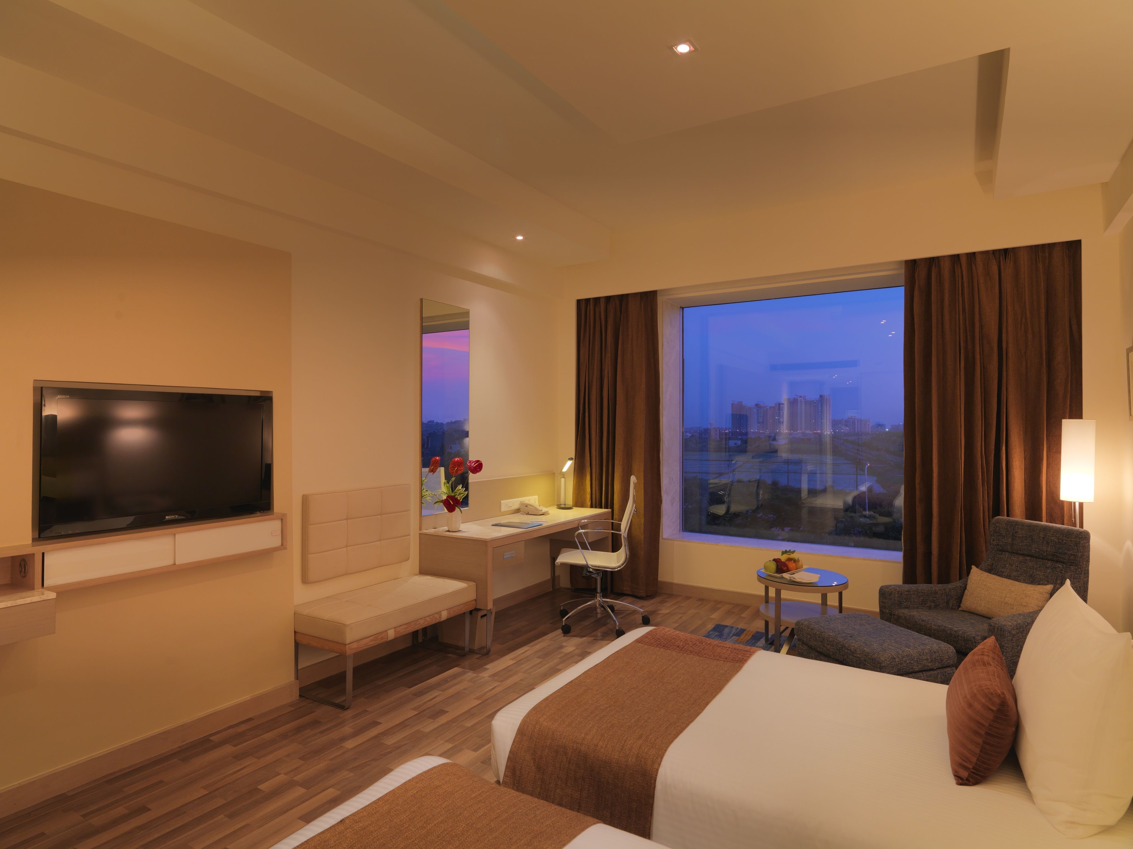 A hotel room with a bed, a desk, a chair, a TV, and a large window with a city view.