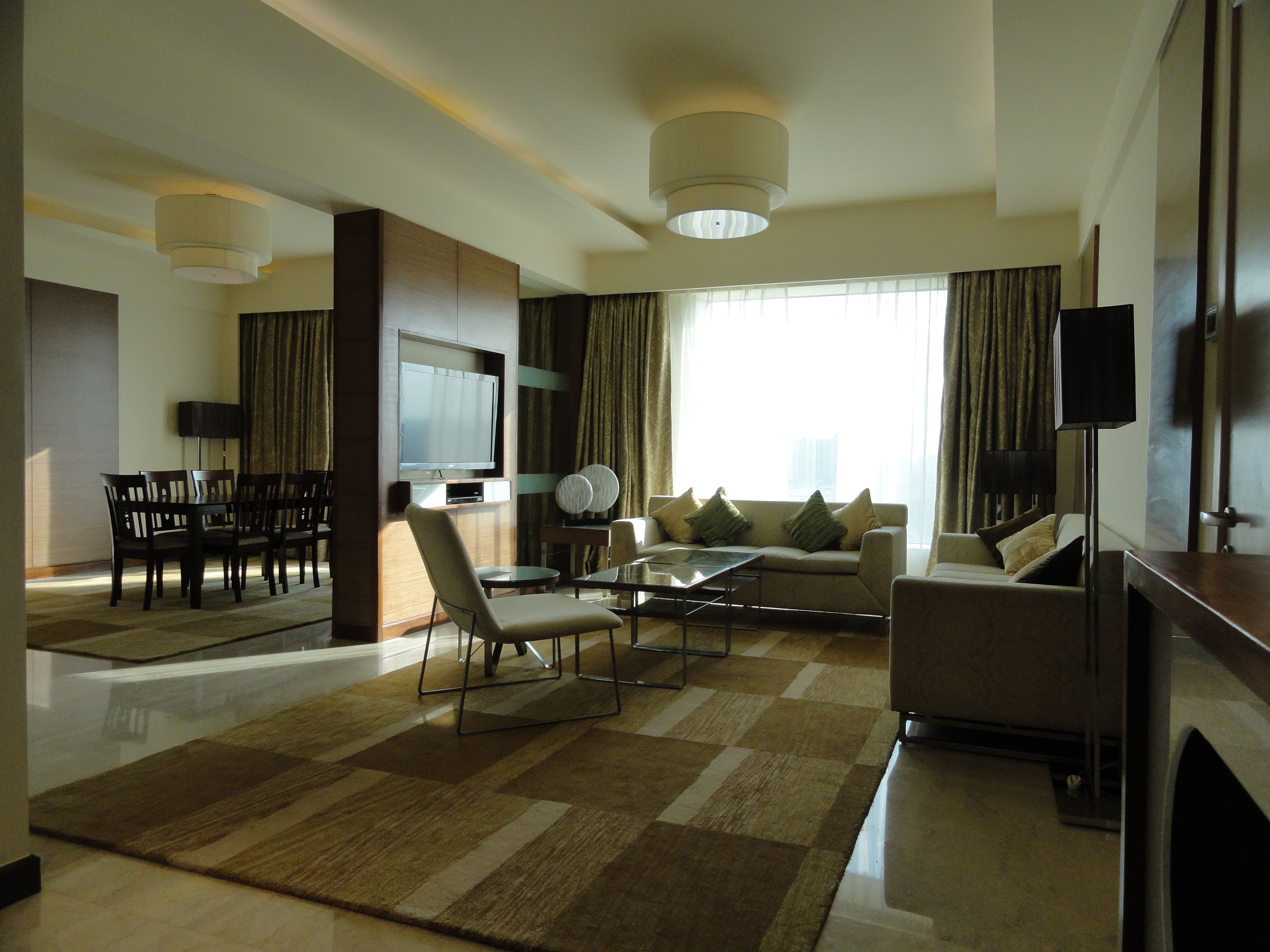 A modern hotel living room with a flat-screen TV, comfortable seating, a coffee table, and a large window with curtains.