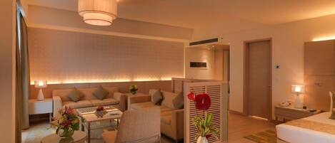 A modern hotel room with a sofa, a dining table, a chair, a bed, and a nightstand with a lamp.