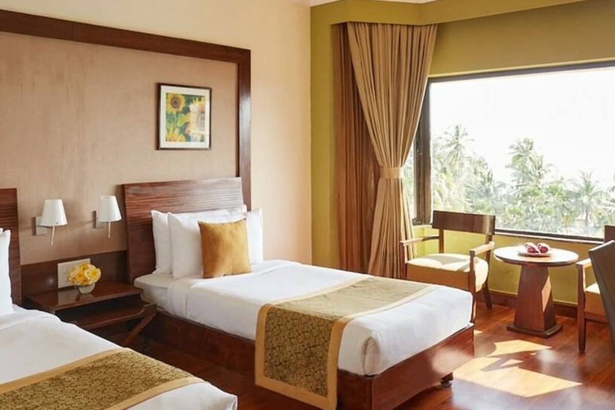 Habitación de hotel con dos camas, una mesita con frutero, una silla y vistas a palmeras a través de amplias ventanas.