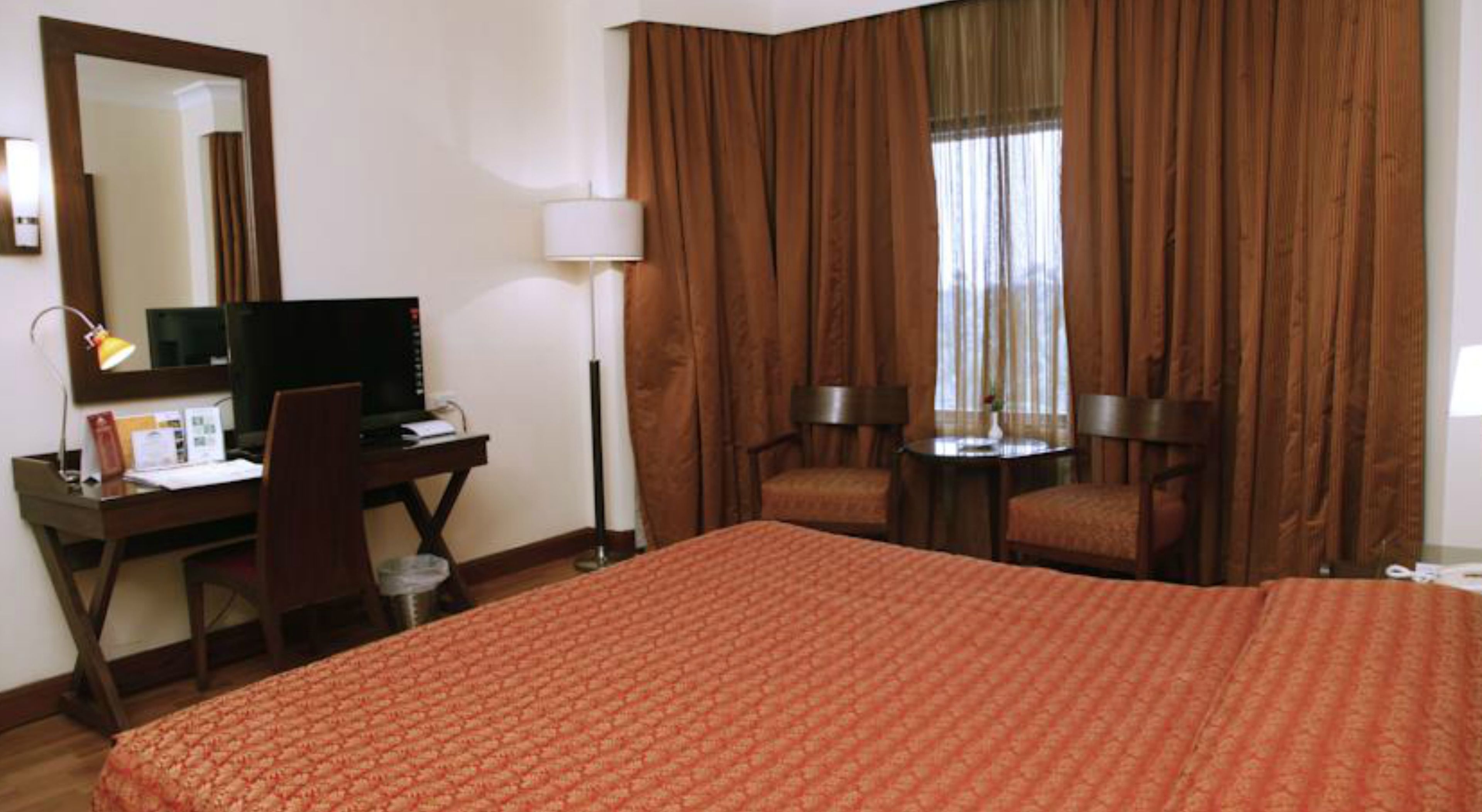 A hotel room with a large bed, a desk with a computer, a chair, a small table, and a lamp.