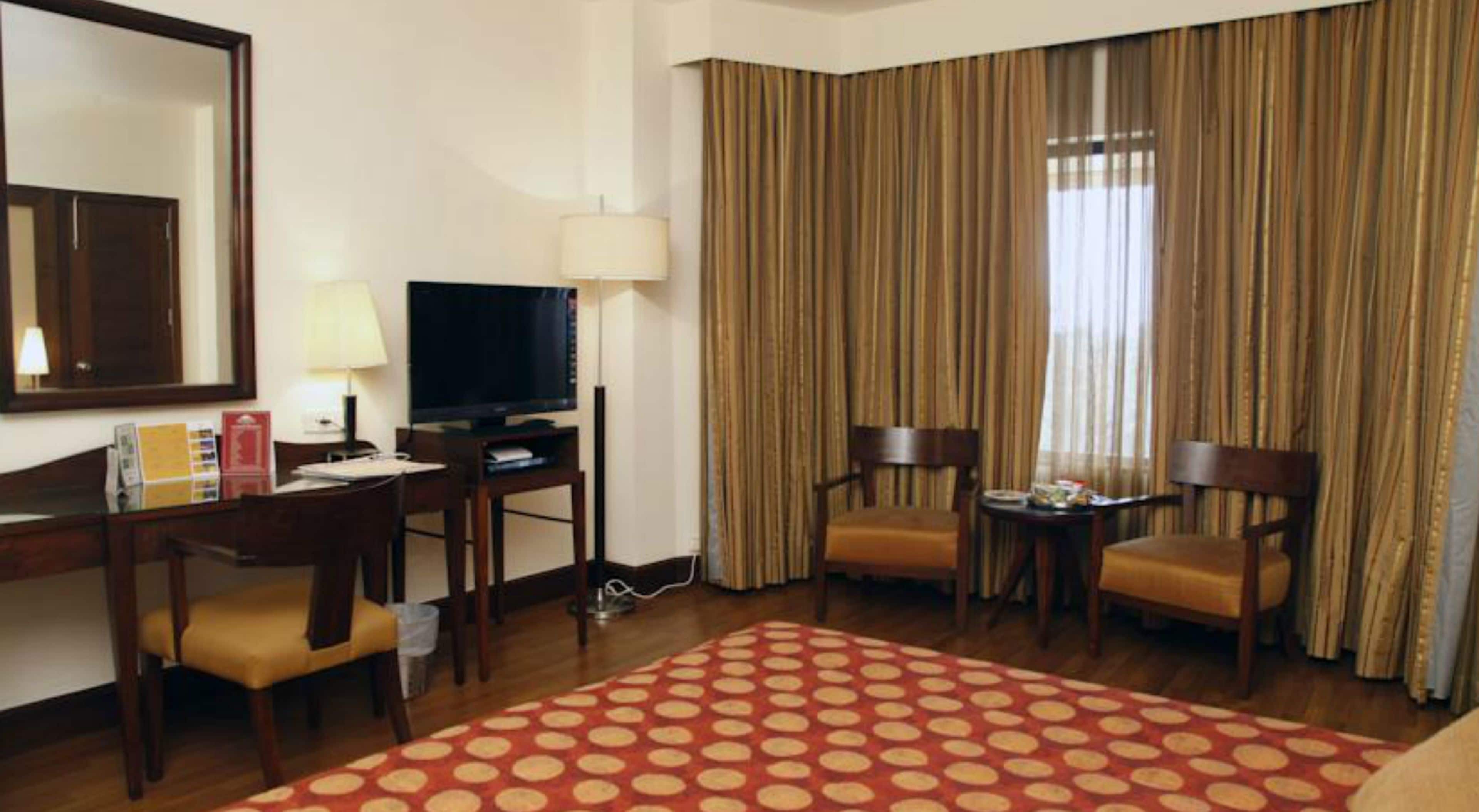 Een hotelkamer met een bureau, twee stoelen, een televisie, een lamp en een groot raam met gordijnen.