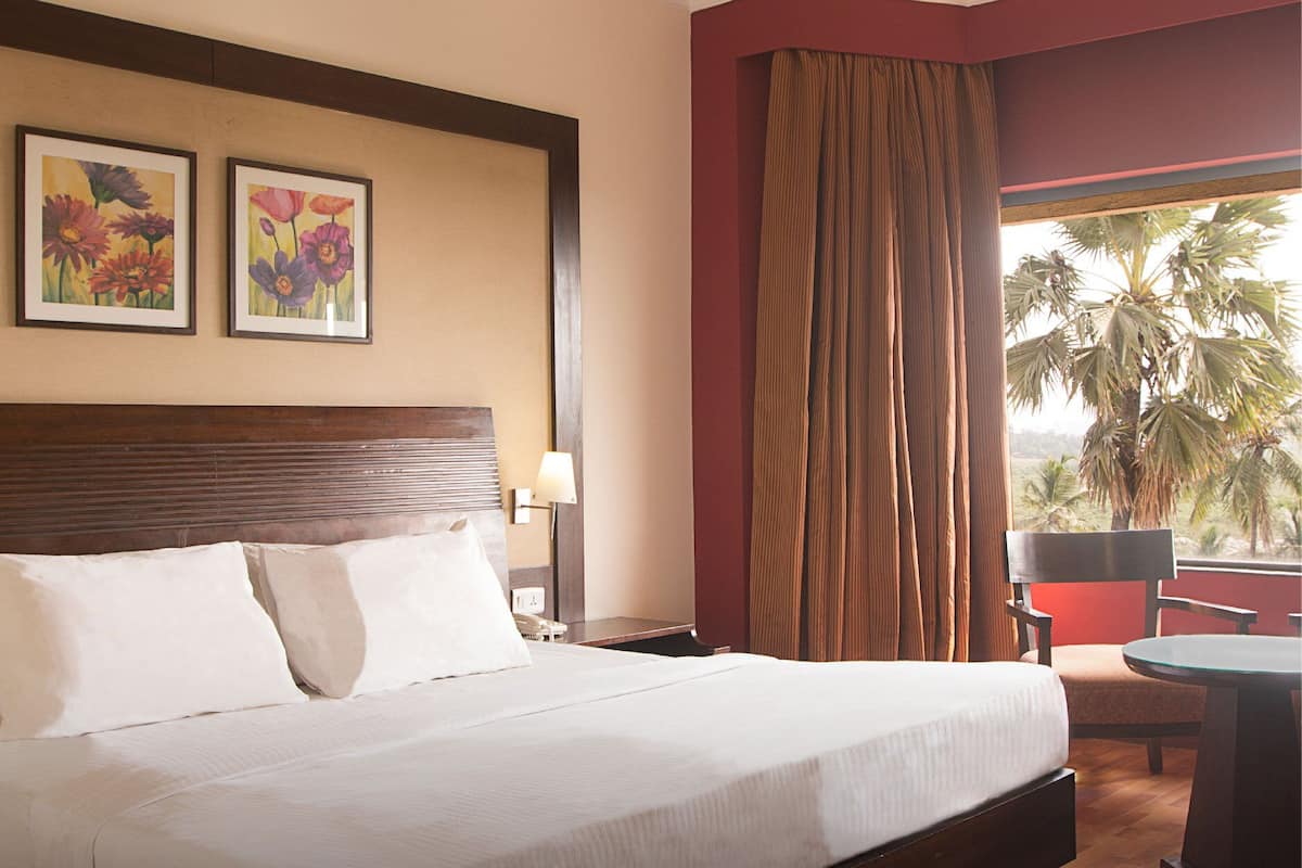 Habitación de hotel con cama, mesita de noche, lámpara, silla, mesa y ventana con cortinas.