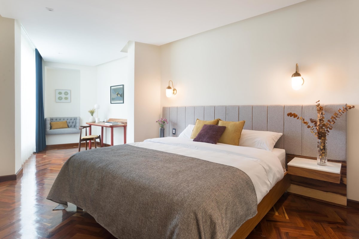 A modern bedroom with a large bed, bedside tables, and a nightstand. There is a seating area with a sofa and a small table.