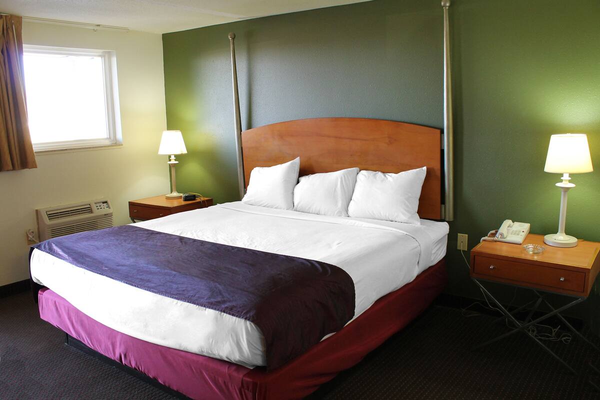A hotel room with a large bed, two bedside tables with lamps, a window with curtains, and a wall-mounted air conditioning unit.