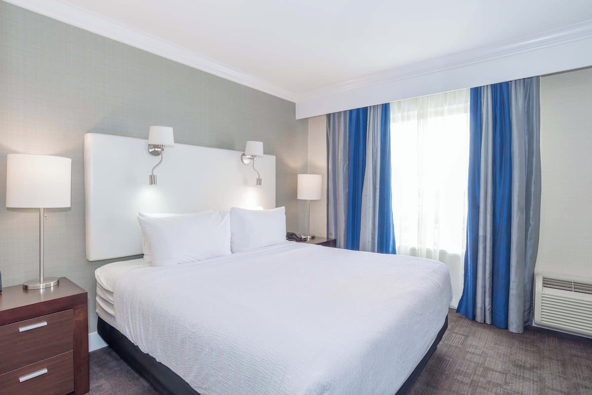 A hotel room with a large bed, two bedside tables with lamps, a nightstand, and a window with blue curtains.