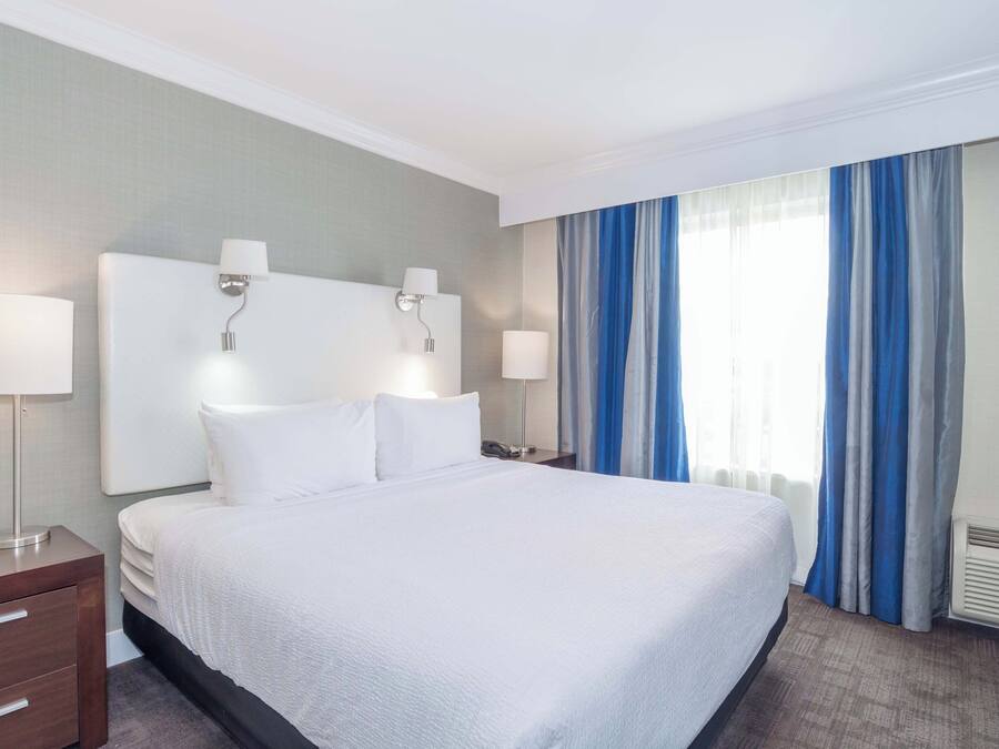 Una habitación de hotel con una cama grande, dos mesitas de noche con lámparas, un velador y una ventana con cortinas azules.