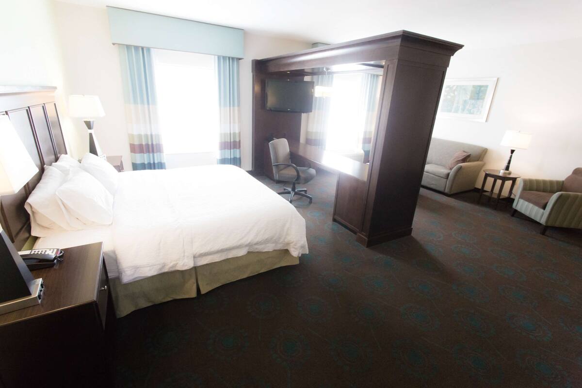 Quarto de hotel com uma cama grande, uma escrivaninha com telefone, televisão, cadeira, mesa pequena com abajur e um sofá.