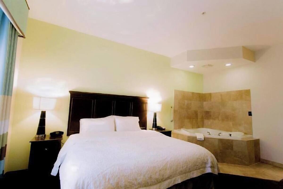 Quarto de hotel com cama grande, banheiro separado com banheira e mesa de cabeceira com luminária.