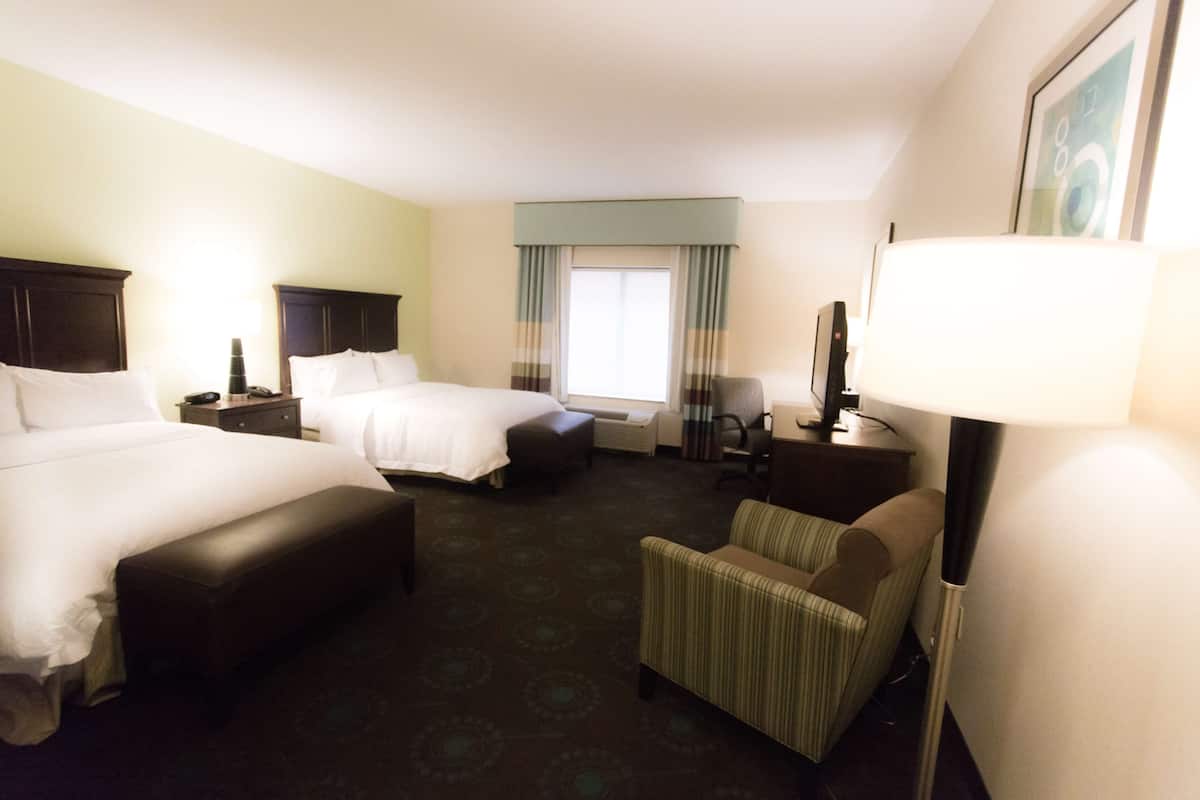 Quarto de hotel com duas camas, uma cadeira, uma escrivaninha, um abajur e uma janela com cortinas.