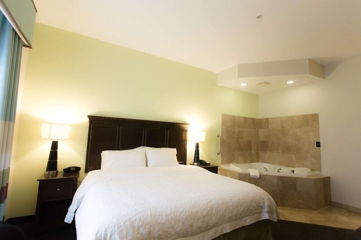 A hotel room with a bed, bedside lamps, a nightstand, a large mirror, and a bathtub.