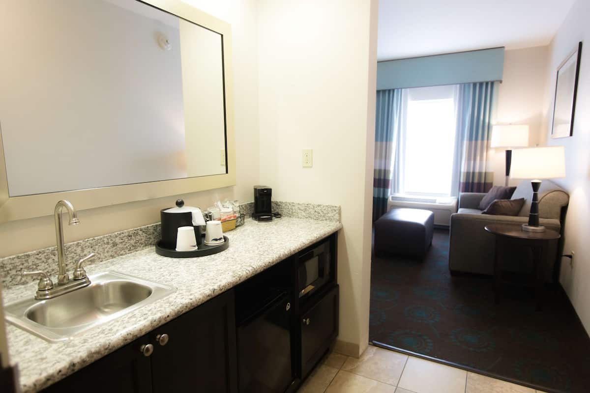 Quarto de hotel com cozinha equipada, incluindo pia, bancada e cafeteira. Possui área de estar com sofá e uma pequena mesa.