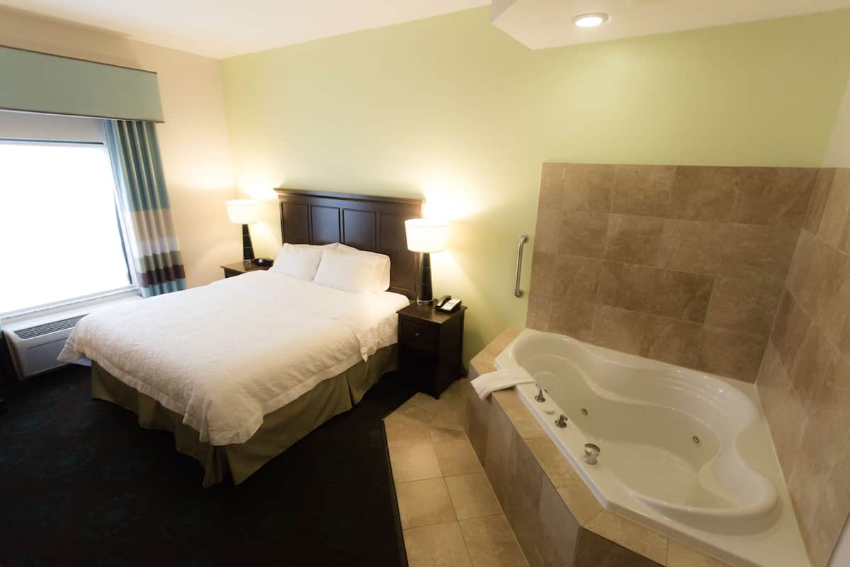 Quarto de hotel com uma cama grande, abajures, banheira e uma janela com cortinas.