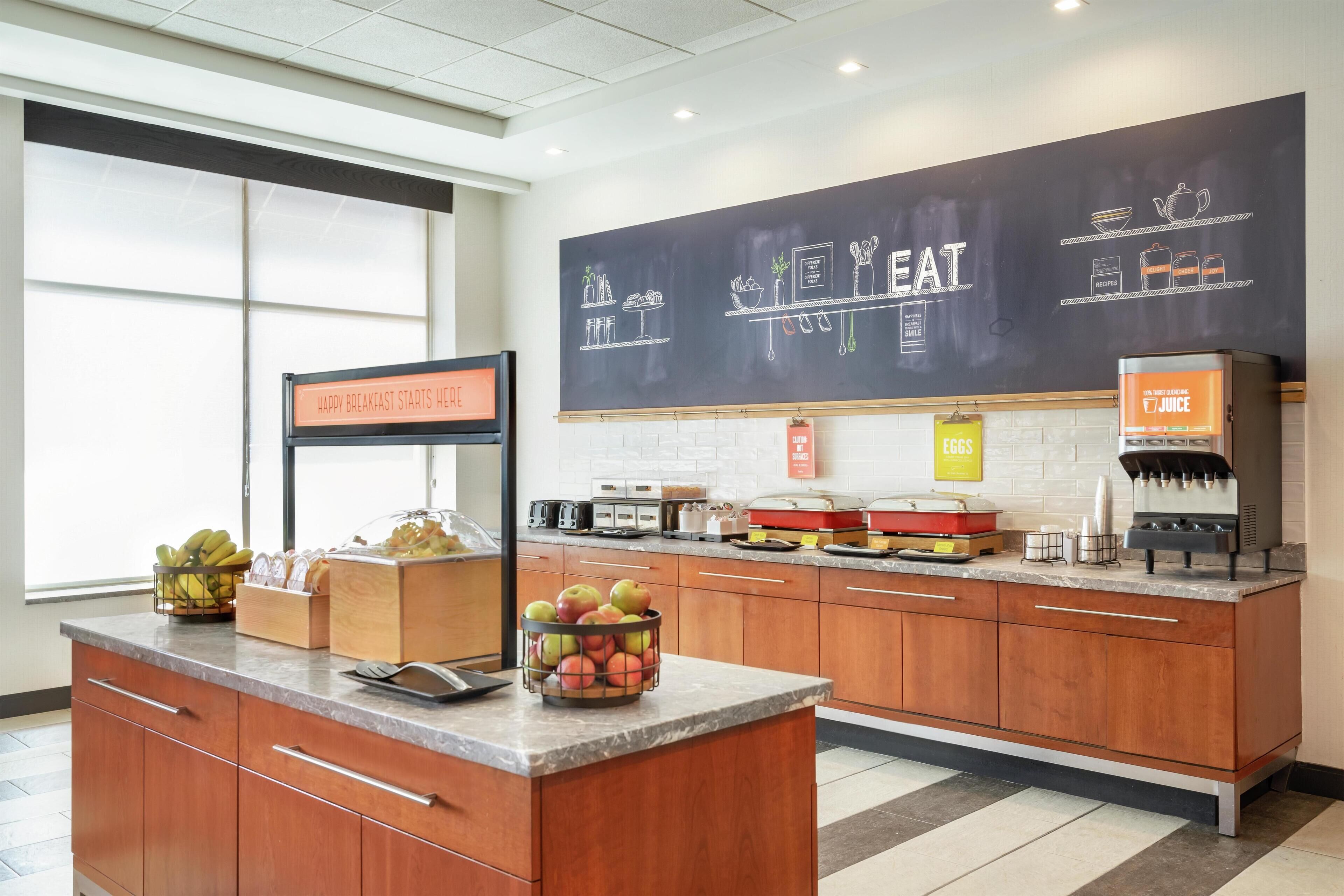 A modern kitchen with a chalkboard displaying food items, a juice dispenser, and a sign indicating breakfast availability.