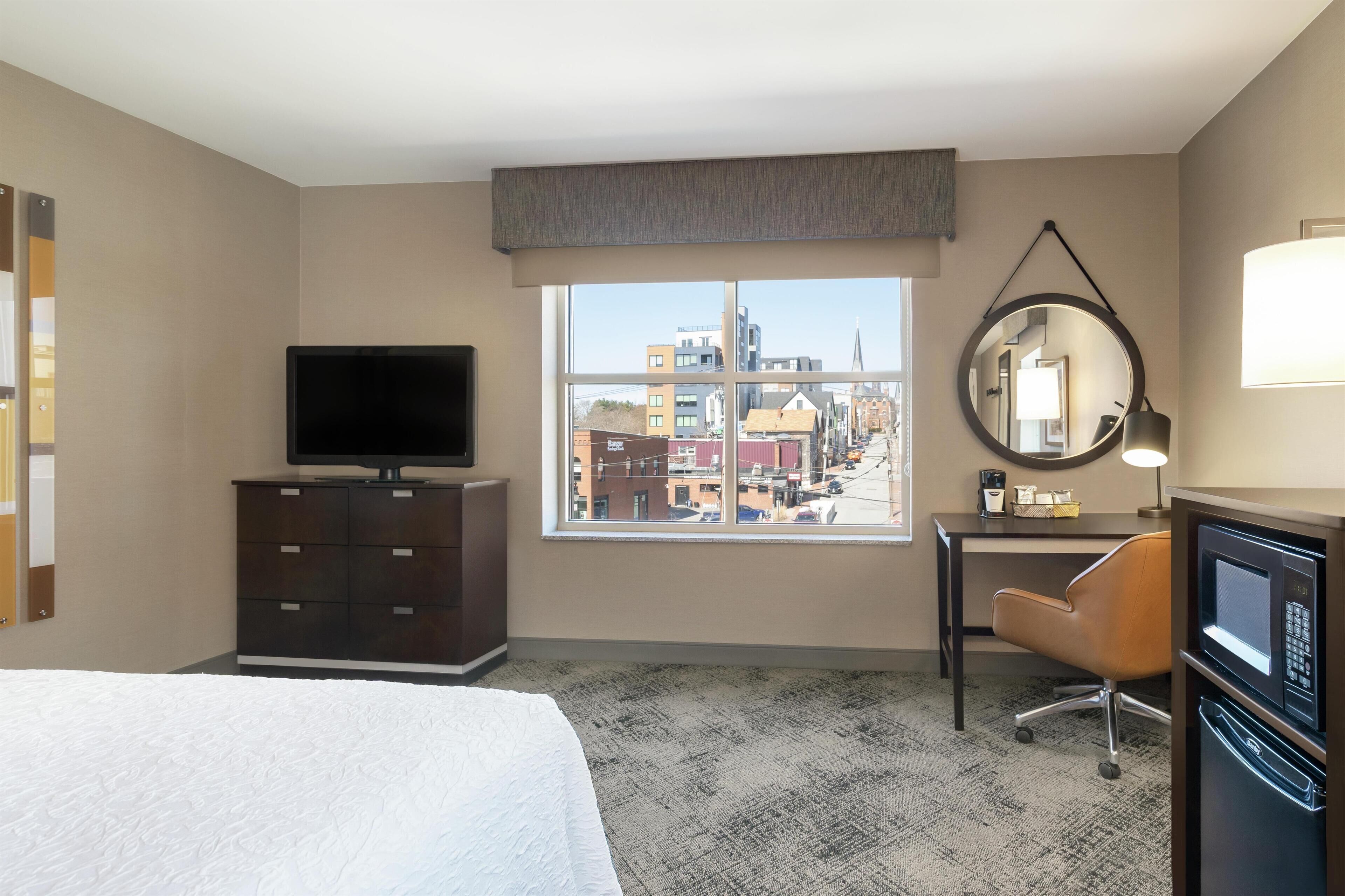 A hotel room with a bed, a desk, a chair, a television, a mirror, and a view of the city through the window.