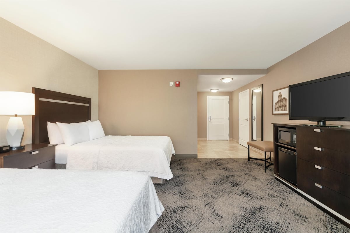 A hotel room with two beds, a desk with a lamp, a dresser, a television, and a door leading to another room.