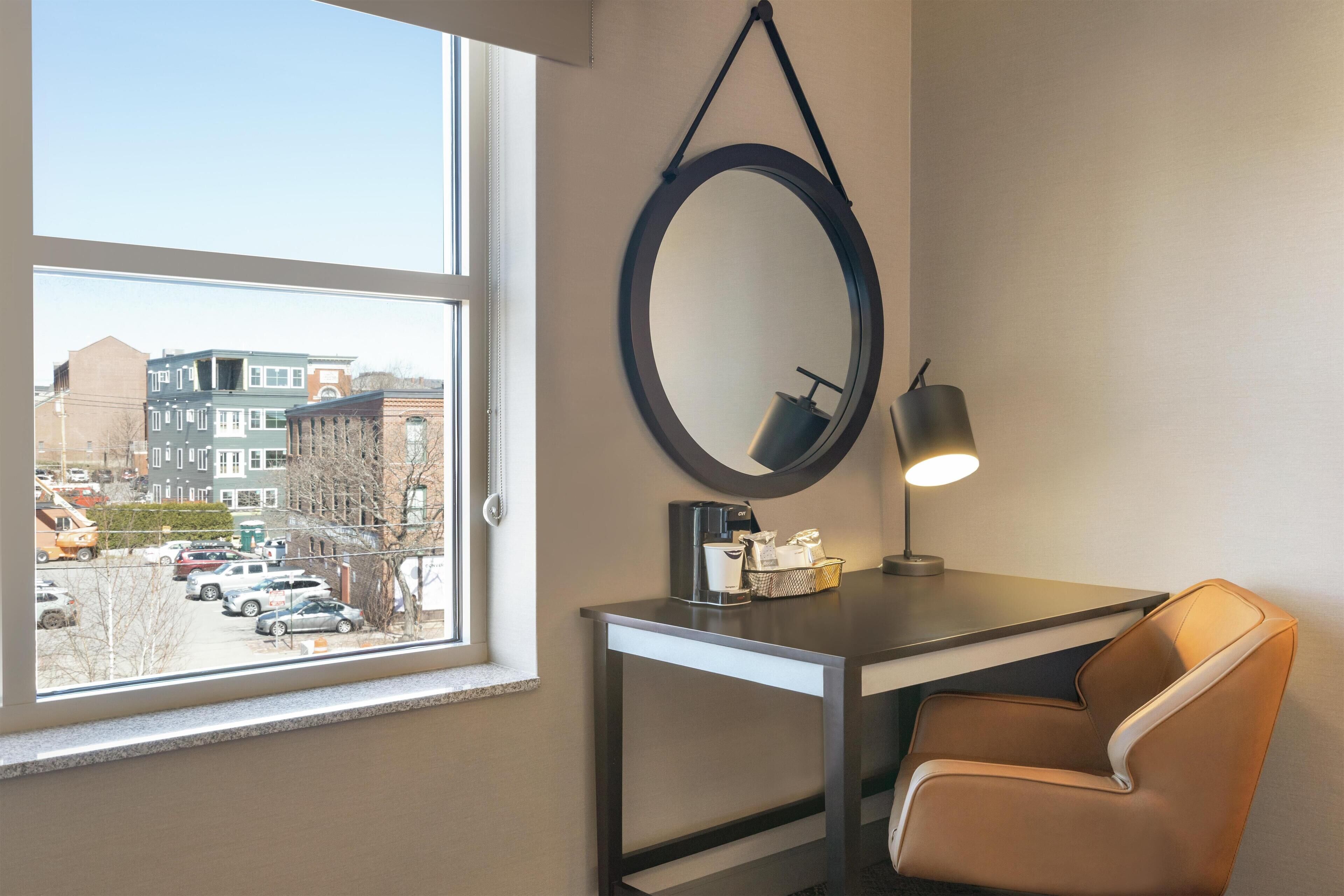 A hotel room with a desk, chair, and a large window offering a view of parked cars and buildings outside.