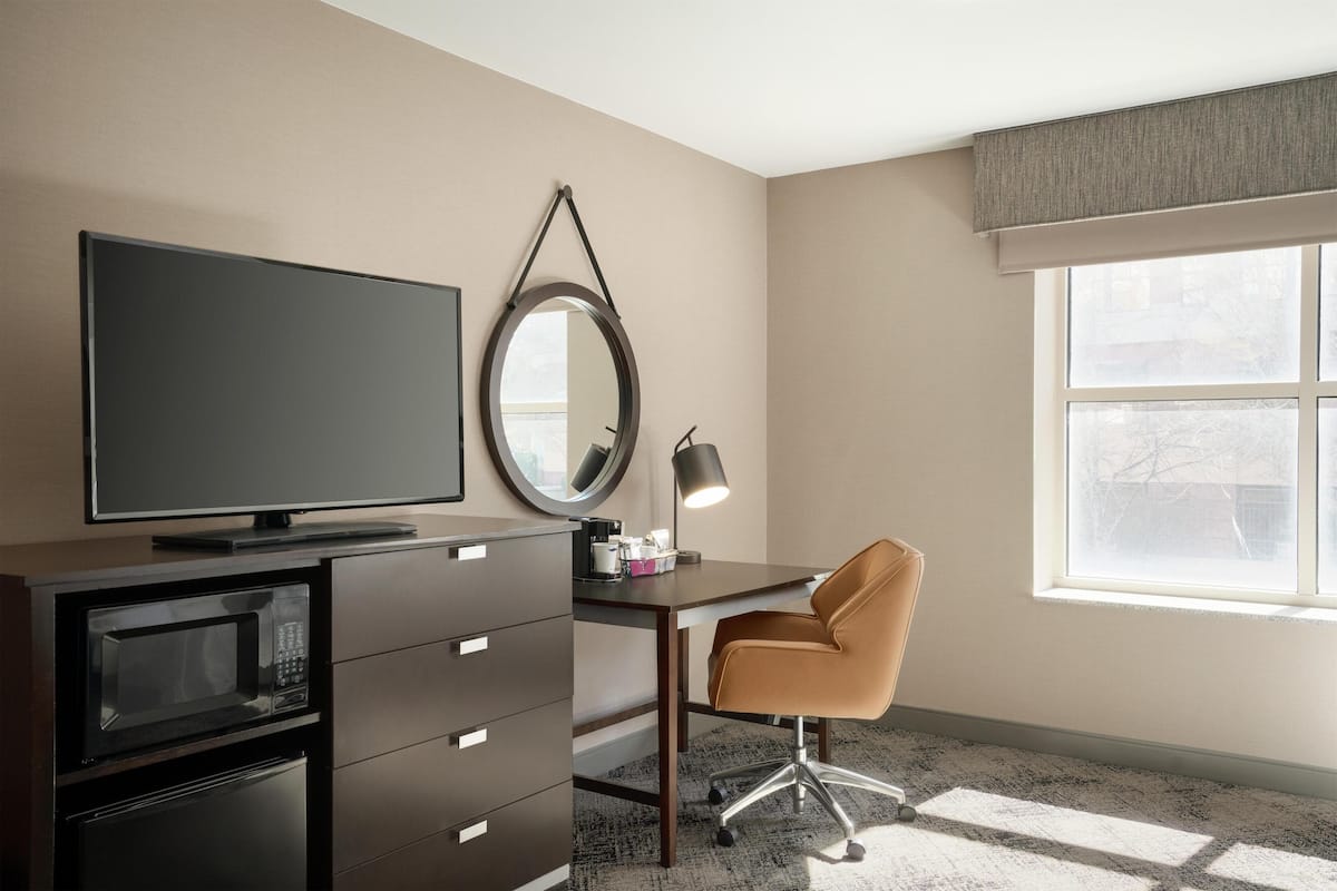 A modern hotel room with a flat-screen TV, a desk with a chair, a mirror, and a large window.