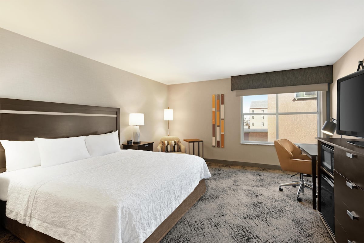 A hotel room with a large bed, a desk, a chair, a television, and a window with a view of buildings.