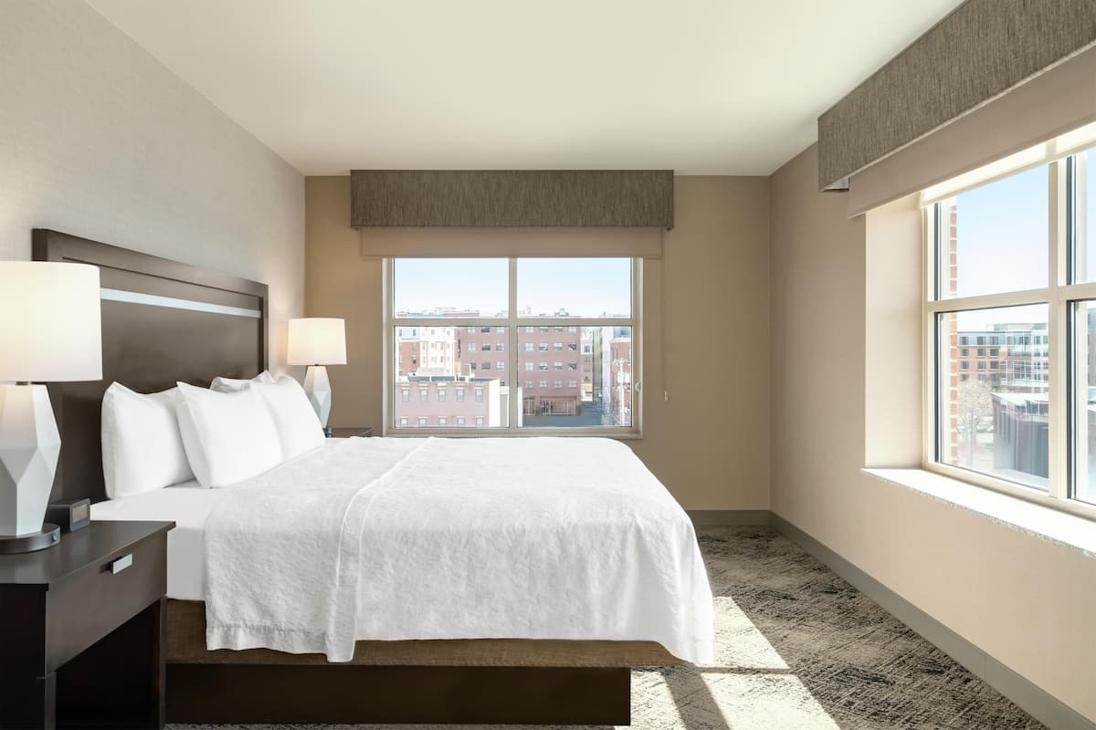 A hotel room with a large bed, two bedside lamps, a nightstand, and a window with a city view.