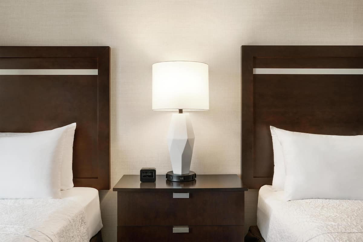 A hotel room with two beds, a nightstand with a lamp, and a small clock.