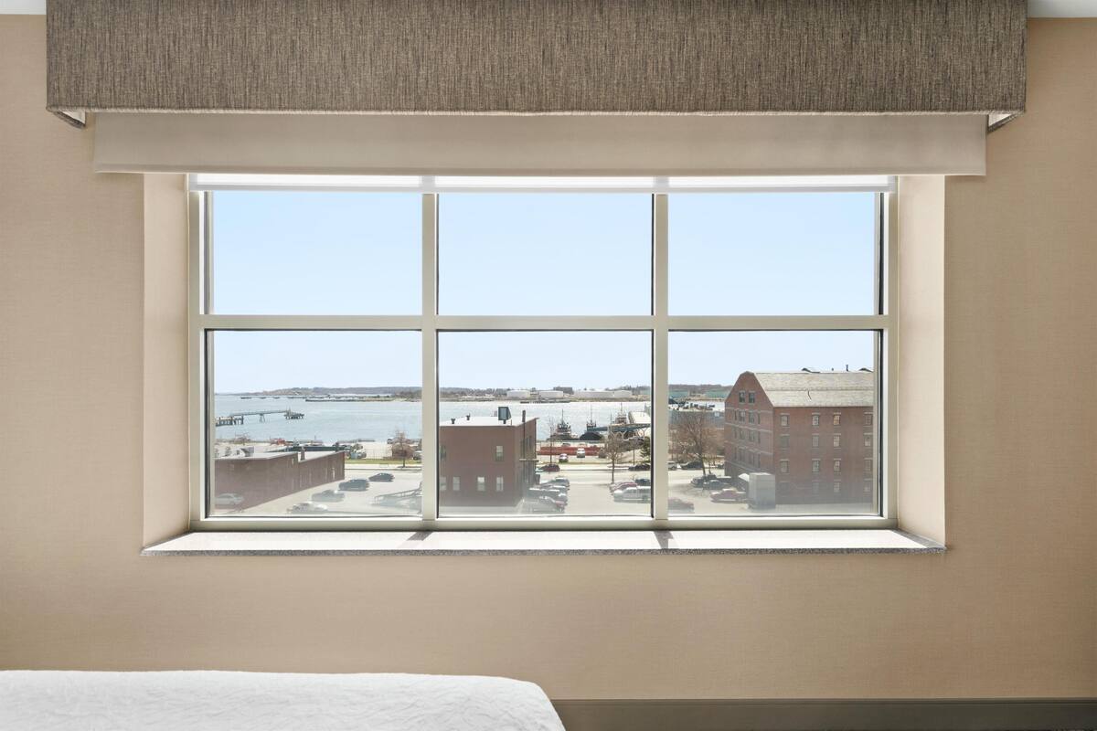 A hotel room with a large window offering a view of a waterfront and buildings.