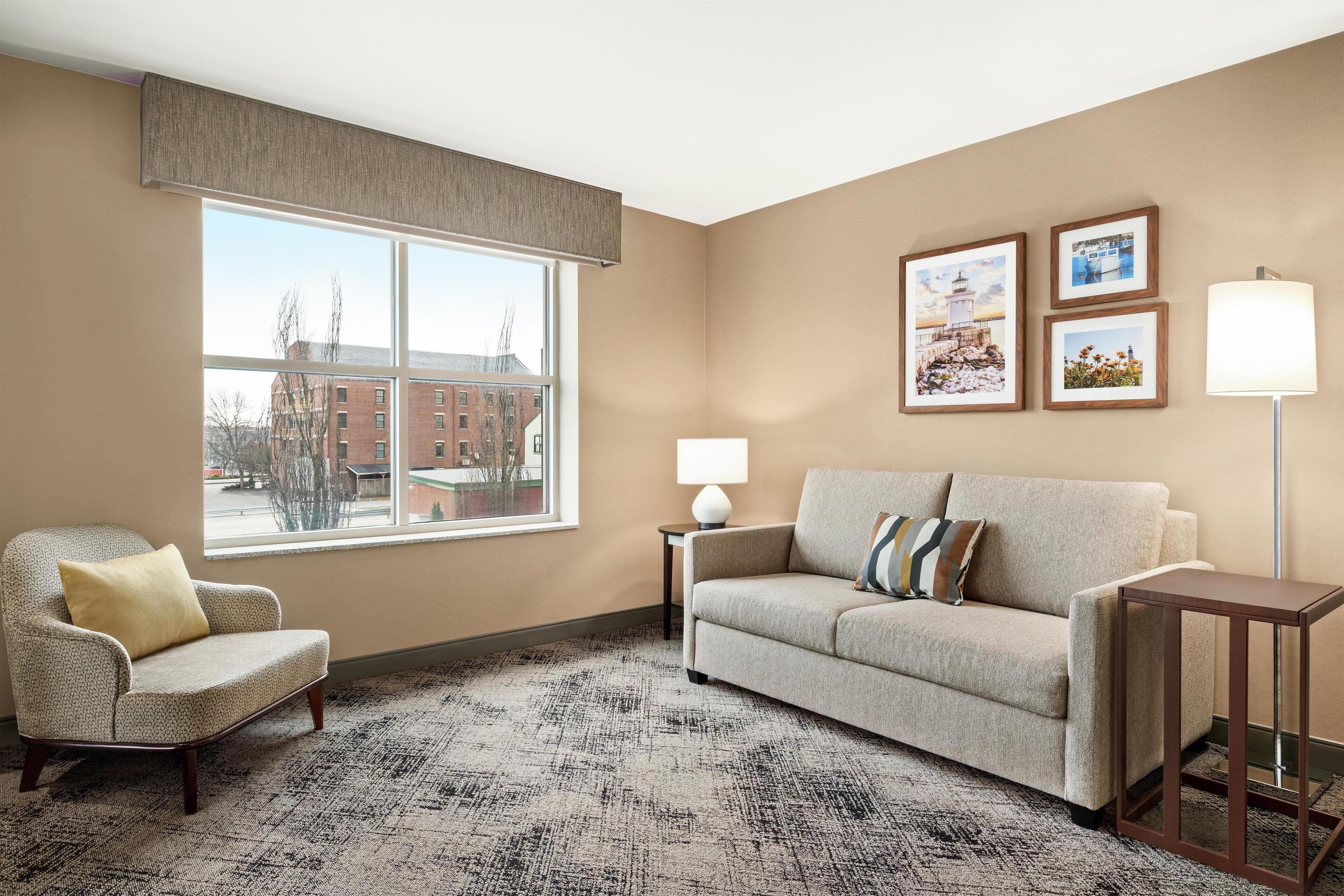 A living room with a beige armchair, a sofa, a small table, and a floor lamp. There are three framed pictures on the wall.