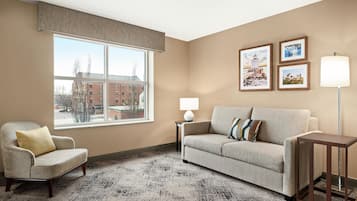 A living room with a beige armchair, a sofa, a small table, and a floor lamp. There are three framed pictures on the wall.
